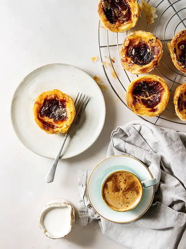 Traditionelles portugiesisches Eierkuchen-Dessert Pasteis Pastel de nata auf einem Kühlgitter und einem Keramikteller mit Gabel, einer Tasse schwarzem Kaffee und einem Kännchen Sahne vor einem weißen Marmorhintergrund.