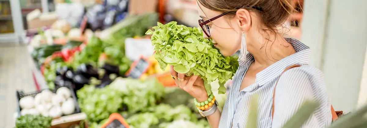 Junge Frau, die einen frischen Salat auswählt, steht mit einem Korb auf dem Lebensmittelmarkt 